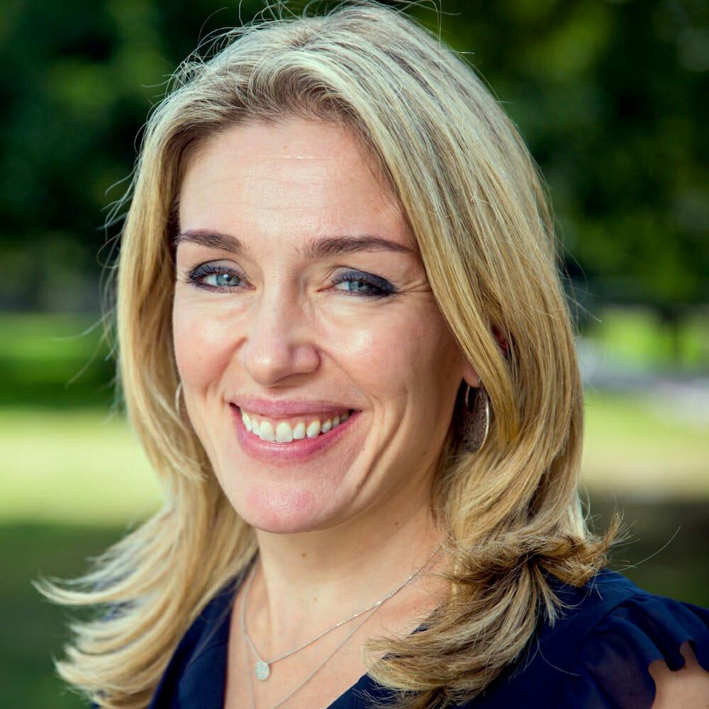 A headshot of Abigail Marsh, smiling in an outdoor setting. She has blonde hair and blue eyes and is wearing a blue shirt.