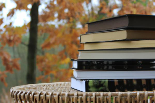A photograph of a pile of fall books sitting on a wicker table with autumn trees in the background