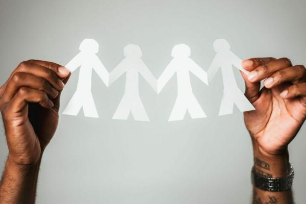 Image of two hands in front of a light gray background holding up a paper chain of people that are not experiencing loneliness because they are holding hands.