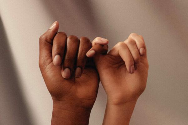 Two hands linking pinkies with a darker skinned hand on the left and a lighter skinned hand on the right in front of a coffee-colored background