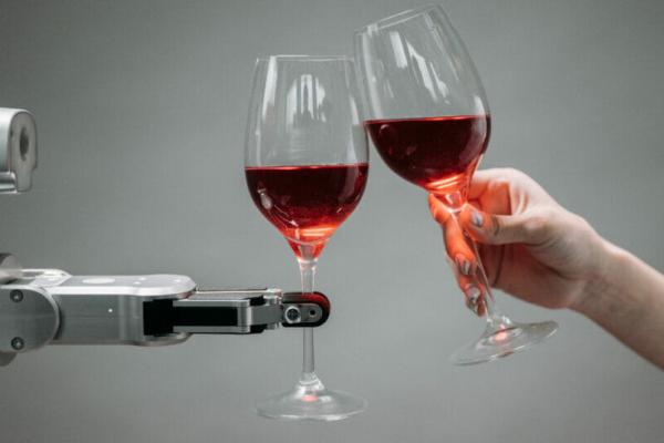 A robot hand holding a wineglass and clinking glasses with a human hand that is also holding a wineglass in front of a medium gray background