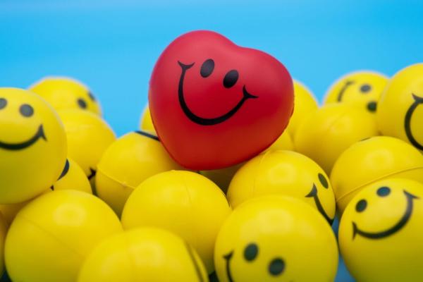 A group of yellow stress balls with happy faces on them in front of a bright blue background. There is one bright red ball with a happy face on top of the yellow ones.