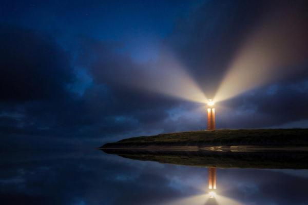 A lighthouse on a jetty sticking out into a body of water, showing light beams, implying hope and resilience