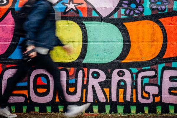 A man in blue pants and a dark blue jacket walking quickly in front of colorful graffiti that says "courage."