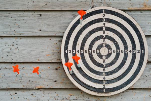 An image of a target with a bullseye target against a plain wooden wall. The dartboard has darts all aound it but none hit the target, implying failure