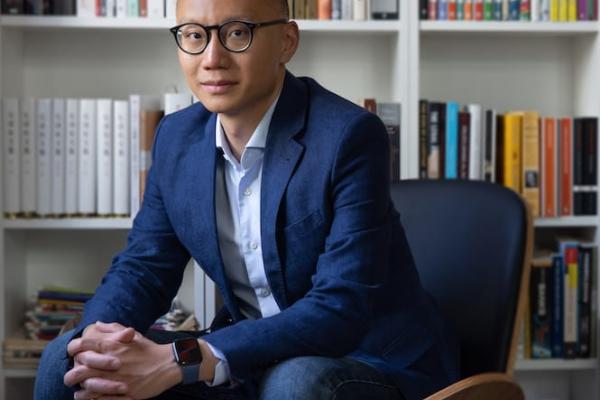 An image of Dan Wang in a blue suit sitting in a brown chair in front of a white bookshelf filled with colorful books. He has short hair, dark rimmed glasses and a lovely watch.